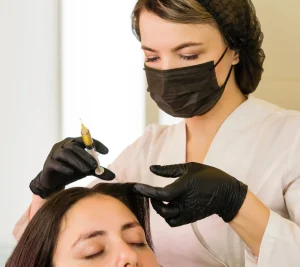 A professional applies a scalp treatment to a client, both wearing black masks and gloves.