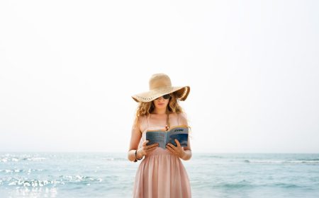 A woman reading a book while standing, wearing a straw hat, with the sea in the background.